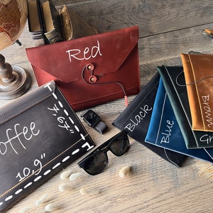 May include: A variety of leather laptop sleeves in different colors, including red, black, blue, green, brown, and coffee. The sleeves are all made of high-quality leather and have a simple, elegant design. Each sleeve has a different color and a handwritten label with the color name.