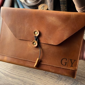 May include: A brown leather envelope with a flap closure and a black cord with brass button closures. The envelope has the initials "G|Y" embossed on the front.