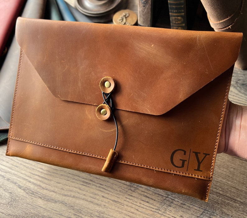 May include: A brown leather envelope with a flap closure and a black cord with brass button closures. The envelope has the initials "G|Y" embossed on the front.
