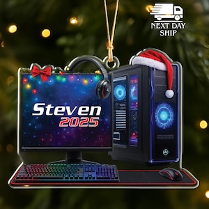 May include: A gaming-themed Christmas ornament featuring a computer monitor, tower, keyboard, mouse, and headset. The monitor displays "Steven 2025" with a festive bow and string lights. A Santa hat rests on the computer tower.