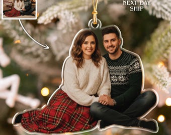 May include: Clear acrylic Christmas ornament featuring a couple. The woman wears a white sweater and a red plaid skirt. The man wears a green sweater with a white pattern and blue jeans. A gold string is attached for hanging. The image in the ornament is of the couple in front of a Christmas tree.