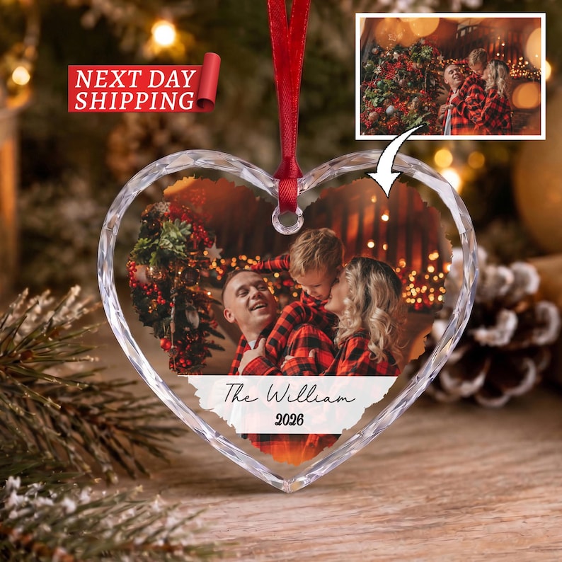 May include: A heart-shaped clear acrylic ornament featuring a printed photograph of a family wearing red and black plaid shirts. The ornament includes a red ribbon for hanging and white text at the bottom that reads The William 2026.