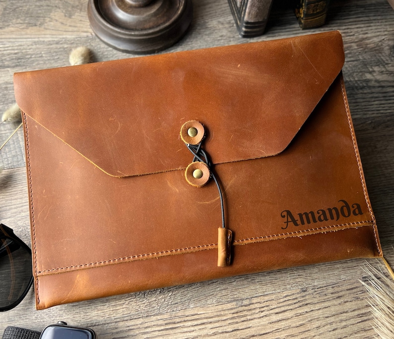 May include: A brown leather envelope with a flap closure and a black cord tie. The envelope has a brass button closure and is personalized with the name "Amanda".