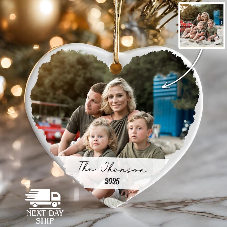 May include: A heart-shaped ceramic Christmas ornament featuring a printed family photograph of two adults and two children. The text "The Thomson" is written in a black script font above the year "2025" in a bold, black serif typeface.