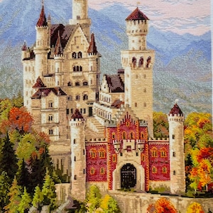 May include: A cross-stitch embroidery of a fairytale castle with a red brick gate and a backdrop of mountains and a blue sky with pink clouds. The castle is surrounded by trees with green and orange leaves.