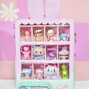 May include: A pastel pink and mint green display backpack featuring two rabbit ears at the top. The front has a clear window divided into twelve compartments, each housing a small collectible figurine. A smaller clear zippered pocket is at the base.