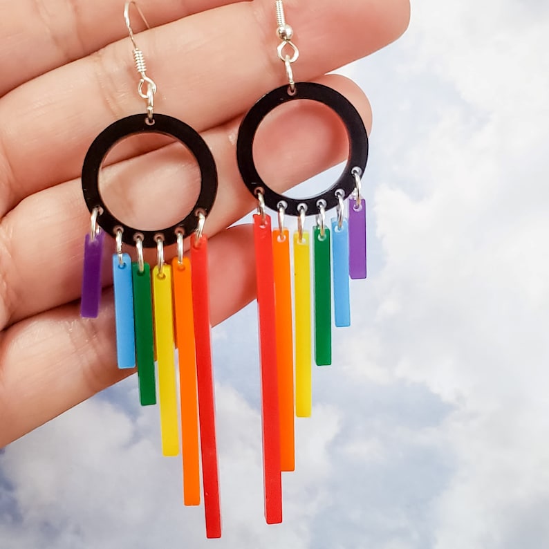 Pu&ograve; includere: Orecchini pendenti arcobaleno con cerchi neri e ganci d'argento. Gli orecchini hanno strisce acriliche color arcobaleno che pendono dai cerchi.