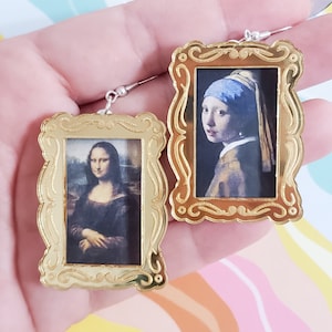 May include: A pair of gold-colored earrings with a picture of the Mona Lisa and Girl with a Pearl Earring in the center. The earrings are in the shape of a picture frame.
