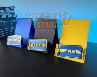 Könnte beinhalten: Drei Präsentationsständer in Blau, Schwarz und Gelb, die jeweils ein "NOW PLAYING"-Schild halten. Die Schilder sind blau mit gelben Rändern und Text. Eine Filmklappe ist im Hintergrund zu sehen.