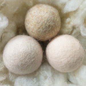 K&ouml;nnte beinhalten: Drei Woll-Trocknerb&auml;lle in einem Nest aus wei&szlig;en Wollfasern. Ein Ball ist beige, die anderen beiden sind hellrosa.
