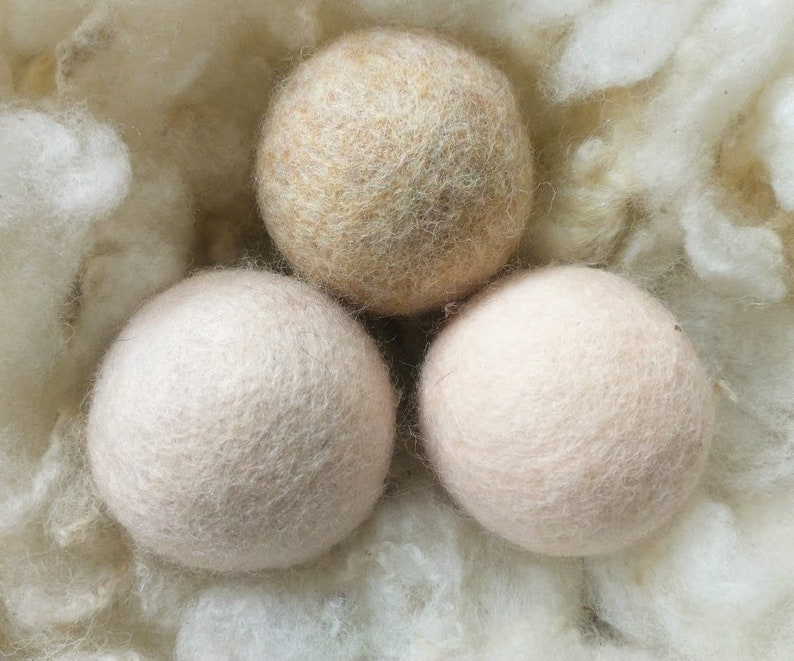 K&ouml;nnte beinhalten: Drei Woll-Trocknerb&auml;lle in einem Nest aus wei&szlig;en Wollfasern. Ein Ball ist beige, die anderen beiden sind hellrosa.