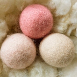 K&ouml;nnte beinhalten: Drei Woll-Trocknerb&auml;lle in einem Nest aus wei&szlig;em Flaum. Ein Ball ist rosa, die anderen beiden sind hellbeige.