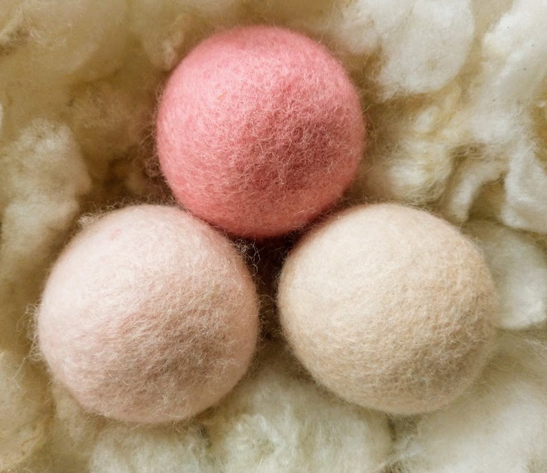 K&ouml;nnte beinhalten: Drei Woll-Trocknerb&auml;lle in einem Nest aus wei&szlig;em Flaum. Ein Ball ist rosa, die anderen beiden sind hellbeige.