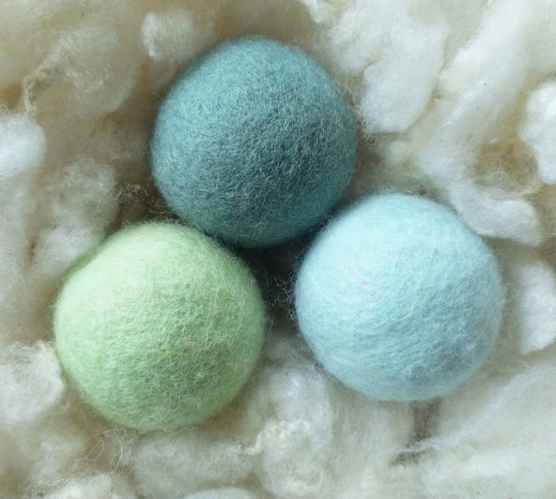 K&ouml;nnte beinhalten: Drei gefilzte Wollkugeln in einem Nest aus wei&szlig;em, flauschigem Material. Die Kugeln sind hellblau, gr&uuml;n und ein hellerer Gr&uuml;nton.