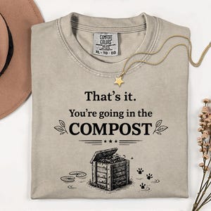 Könnte beinhalten: Ein hellbraunes Comfort Colors T-Shirt mit dem Text "That's it. You're going in the COMPOST" in Schwarz. Das Design umfasst eine Kompostbehälter-Illustration, Pfotenabdrücke und eine goldene Sternkette. Das Shirt ist in Größe XL.