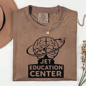 Puede incluir: Camiseta marrón Comfort Colors con un gráfico negro de un cerebro con un avión que lo rodea y el texto "JET EDUCATION CENTER". Un collar dorado con un colgante de estrella descansa sobre la camisa.