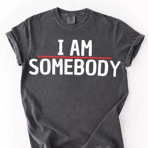 May include: Dark grey t-shirt featuring the white text "I AM SOMEBODY" in a bold font, with a red line underneath "I AM". The shirt has rolled-up sleeves and a crew neck.