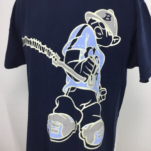 Könnte beinhalten: Ein marineblaues T-Shirt mit einer Comicfigur, die einen blau-weißen Hut trägt und ein Seil hält. Die Figur ist weiß umrandet und trägt ein blaues Hemd und eine blaue Hose.