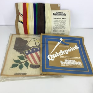May include: A needlepoint kit featuring an eagle with an American flag shield and a green leaf branch. The kit includes yarn, canvas, and instructions. The packaging includes the text "Quickpoint" and "Yarn Brunswick".