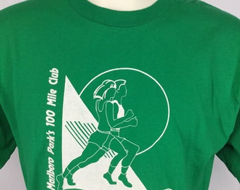 Puede incluir: Camiseta verde con un gráfico blanco de dos personas corriendo. El gráfico incluye el texto "Marlboro Park's 100 Mile Club" y "I take pride in my stride!"
