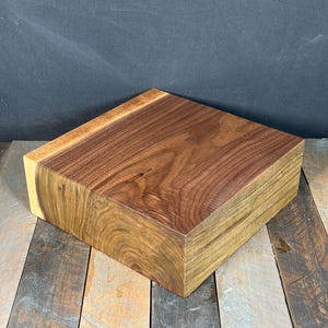 May include: A solid, rectangular wooden block with a natural wood grain pattern. The block is a rich brown color with lighter, yellowish-brown edges. The wood grain is visible on the top and sides.