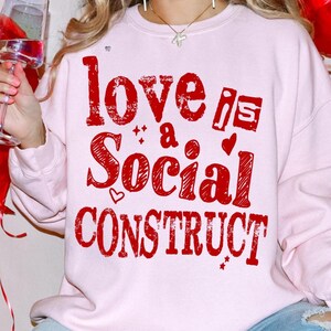 May include: A light pink sweatshirt with the phrase "love is a social construct" in red, distressed lettering. The design includes hearts and stars. The sweatshirt is a casual, everyday garment.