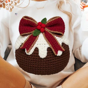 May include: White sweatshirt featuring a crocheted Christmas pudding design. The pudding has a brown base, white icing, and a red bow with gold trim. Green leaves and red berries complete the festive look. A unique, handmade holiday sweater.