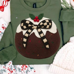 May include: Green sweatshirt featuring a knitted Christmas pudding design. The pudding has a brown base, white icing, and a checkered bow. It is decorated with green leaves and red berries. The sweatshirt has a crew neck and long sleeves.