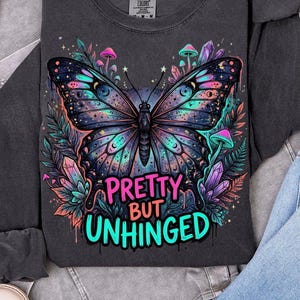 May include: Dark gray long-sleeve shirt featuring a colorful butterfly design with the text "Pretty But Unhinged". The butterfly has blue, pink, and purple accents. The shirt also has mushrooms and crystals.