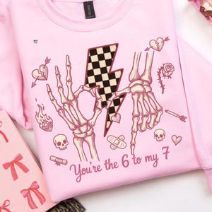 May include: Pale pink sweatshirt with a graphic design. The design includes skeleton hands, a checkered lightning bolt, hearts, and the text "You're the 6 to my 7." There is also a skull, a rose, and a bandage.