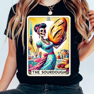 Könnte beinhalten: Schwarzes T-Shirt mit einem Tarotkarten-Design. Die Karte zeigt einen Skelettbäcker, der ein Messer und ein Brotlaib hält. Der Bäcker trägt ein blaues Polka-Dot-Kleid und ein rosa Kopftuch. Der Text "THE SOURDOUGH" steht unten.