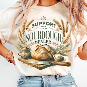 May include: A white t-shirt with a graphic design featuring a loaf of sourdough bread, a jar of jam, a knife, and a small dish of butter. The graphic also includes the text "Support Your Local Sourdough Dealer".