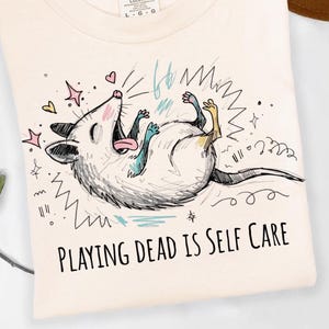May include: Cream-colored sweatshirt featuring a playful illustration of a cartoon opossum lying on its back. The opossum is surrounded by doodles and the text "PLAYING DEAD IS SELF CARE" is printed below the illustration.