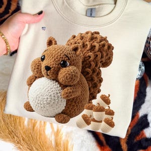 May include: Cream-colored sweatshirt featuring a crocheted squirrel holding a white ball. The squirrel is brown with a fluffy tail and cheeks. Several crocheted acorns are below the squirrel. The sweatshirt is held by a person.