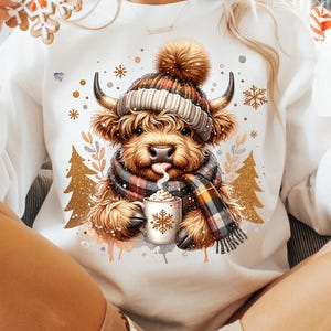 May include: White sweatshirt featuring a Highland cow illustration. The cow is wearing a plaid hat and scarf, holding a mug of hot chocolate. Gold Christmas tree and snowflake accents surround the cow. A festive and cozy design.