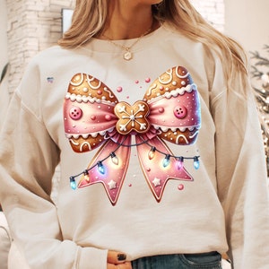 Peut inclure: Un sweat-shirt crème avec un nœud en pain d'épices rose et brun avec des lumières blanches et un flocon de neige.