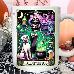 May include: White ceramic mug with a colorful illustration of a skeleton woman with long purple and blue hair, holding a beer mug and a cauldron with green smoke. A white dog stands next to her. The text "HAIR OF THE DOG" is at the bottom of the mug.