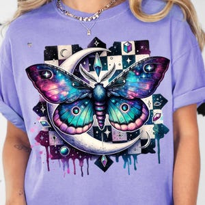 May include: A light purple t-shirt featuring a colorful moth design. The moth has galaxy-themed wings with shades of blue, purple, and pink. The design includes a crescent moon, geometric shapes, and a checkered background.