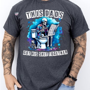 May include: A dark gray t-shirt with a humorous graphic of a skeleton sitting on a toilet reading a newspaper. The text on the shirt reads "This Dad's Got His Shit Together".