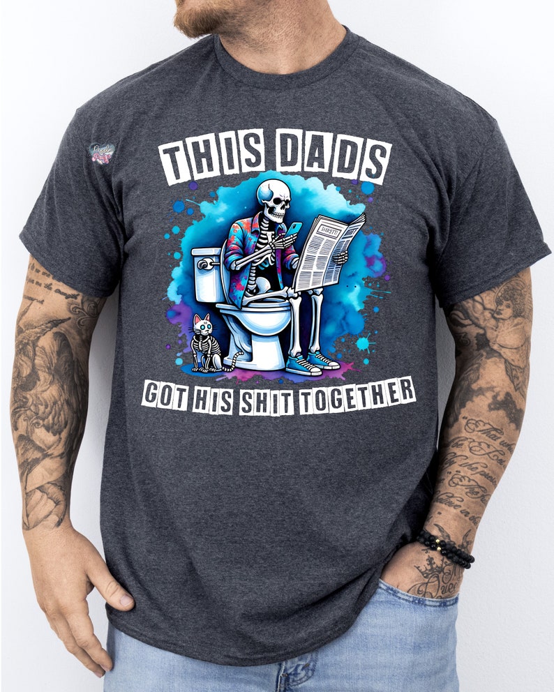 May include: A dark gray t-shirt with a humorous graphic of a skeleton sitting on a toilet reading a newspaper. The text on the shirt reads "This Dad's Got His Shit Together".