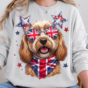 May include: A light gray crewneck sweatshirt featuring a graphic of a golden-brown dog wearing round sunglasses with a Union Jack pattern. The dog wears a matching Union Jack bandana and is surrounded by red, white, and blue star-shaped accents.