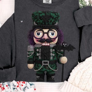 May include: A dark gray sweatshirt featuring a crocheted nutcracker doll. The doll has a green and black hat, purple hair, and a black mustache. It holds a staff with a bat on top. The doll is wearing a green and black uniform.