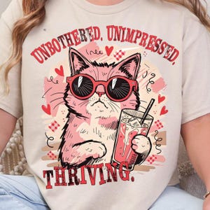 May include: Beige t-shirt featuring a pink cat wearing red sunglasses, holding a drink with ice. The shirt has the text "UNBOTHERED, UNIMPRESSED, THRIVING." surrounded by hearts and decorative elements.