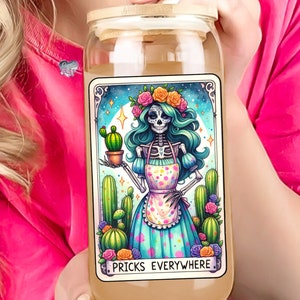 Puede incluir: Un tarro de vidrio con tapa de madera y una ilustraci&oacute;n colorida de un esqueleto con una corona de flores y un delantal de lunares que sostiene un cactus. El texto "Pricks Everywhere" est&aacute; impreso debajo de la ilustraci&oacute;n.