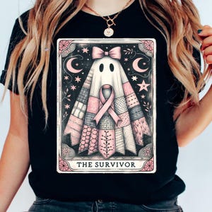 May include: Black t-shirt featuring a graphic of a ghost with a pink ribbon, bow, and patchwork design. The image includes the text "THE SURVIVOR" at the bottom, with stars and crescent moons in the background. The design is framed with a pink border.