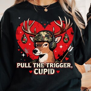 May include: Black sweatshirt with a deer graphic wearing a camouflage helmet and headphones, set against a red heart. The deer has antlers and is flanked by two grenade-shaped ornaments. Text reads "PULL THE TRIGGER, CUPID" with hearts.