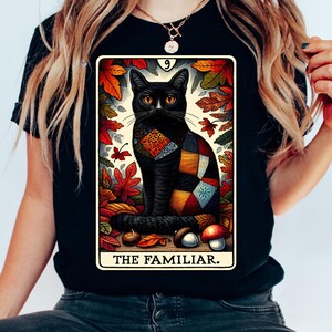 Op de afbeelding: Zwart T-shirt met een tarotkaartontwerp. De kaart toont een zwarte kat met oranje ogen, gekleed in een patchwork quilt, omringd door herfstbladeren. De tekst "THE FAMILIAR" staat onderaan de kaart.