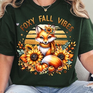 May include: A green t-shirt with the text "Foxy Fall Vibes" in gold glitter lettering. A cartoon fox with a scarf and a flower crown is surrounded by sunflowers, pumpkins, and fall leaves.