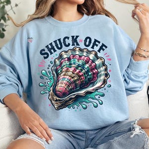 May include: Light blue sweatshirt with the text "SHUCK*OFF" printed in black. The graphic features a colorful oyster shell with a pearl inside, surrounded by blue and green water droplets.