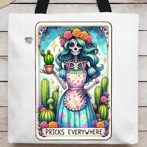 Puede incluir: Una bolsa de tela blanca con una ilustraci&oacute;n colorida de un esqueleto con una corona de flores y un vestido azul y blanco sosteniendo un cactus. El texto "Pricks Everywhere" est&aacute; impreso debajo de la imagen.